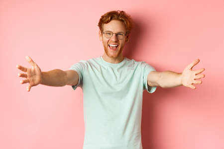 Young friendly man with red hair and beard reaching hands for hug, stretch out arm in warm welcome, smiling happily, standing over pink backgroundの写真素材