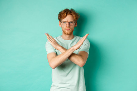 Serious and confident redhead man in t-shirt and glasses saying no, showing cross gesture to stop you, refucing or declining something, standing over turquoise backgroundの写真素材