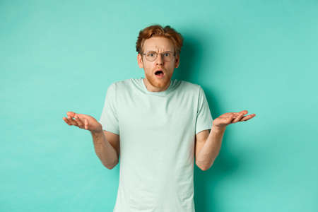 Confused and shocked guy with red messy hair and glasses, shrugging and raising hands, staring at something strange, cant understand, standing over turquoise backgroundの写真素材