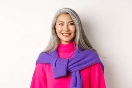 Close-up of beautiful asian senior woman in trendy pink sweater, smiling cheerful at camera, standing over white backgroundの写真素材