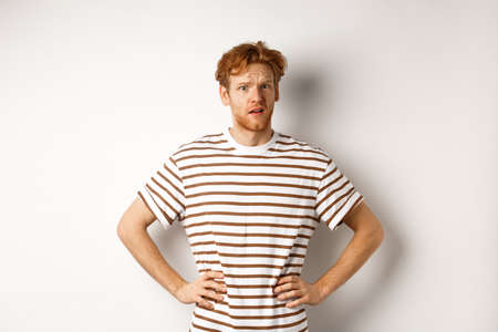 Confused and offended man with ginger curly hair staring at camera puzzled, standing questioned against white backgroundの写真素材