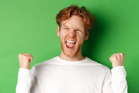 Close-up of lucky young man winner, celebrating victory, shouting yes with joy and making fist pumps, triumphing on green backgroundの写真素材