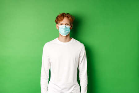 Covid-19, social distancing and lifestyle concept. Young redhead man wearing face mask during coronavirus pandemic, standing over green backgroundの写真素材