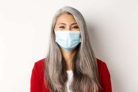 Covid, pandemic and business concept. Close-up of cheerful asian senior businesswoman in medical mask smiling at camera, white backgroundの写真素材