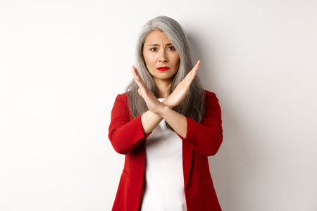 Asian senior woman in red blazer making cross gesture, pleading you stop, disagree and disapprove something, standing over white backgroundの写真素材