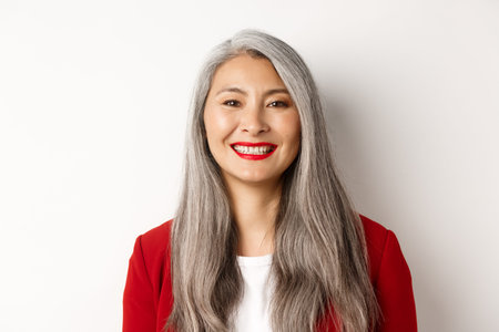 Close-up of elegant mature woman in red blazer and makeup, smiling happy at camera, standing over...