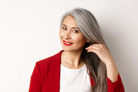 Beauty and haircare concept. Close up of elegant asian senior woman showing shiny and healthy grey hair, smiling and looking aside, white backgroundの写真素材