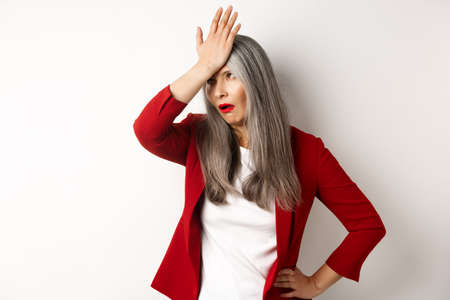 Annoyed asian office lady in red blazer roll eyes and facepalm, standing bothered and irritated over white backgroundの写真素材