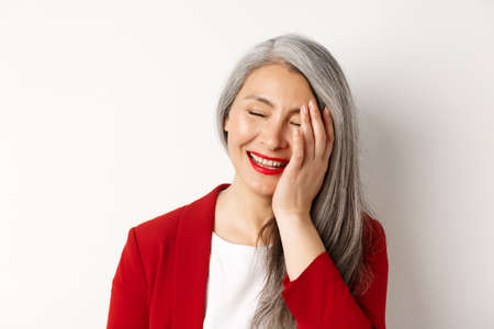 Beauty and makeup concept. Close up of beautiful asian mature woman, laughing and touching face, smiling happy, standing over white backgroundの写真素材
