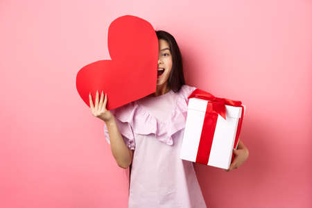 Valentines day concept. Happy young girl asian holding romantic gifts, red heart card from lover and present box, looking amazed at camera, standing in dress on pink backgroundの写真素材