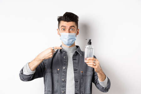 Health and pandemic concept. Stylish guy in medical mask pointing at bottle with hand sanitizer, showing antiseptic, standing on white backgroundの写真素材