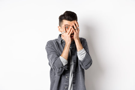 Scared young man peeking through fingers with nervous face, staring at something scary, standing on white backgroundの写真素材