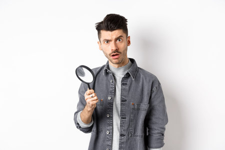 Shocked man found something with magnifying glass, look at camera, standing against white background