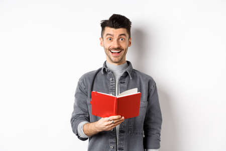 Hobbies and leisure. Happy young man reading planner, holding diary or red journal and smiling, making notes, standing on white backgroundの写真素材