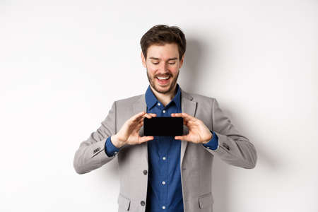 E-commerce and online shopping concept. Happy young man in suit laughing, showing empty smartphone screen horizontally, wite backgroundの写真素材