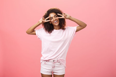 Indoor shot of charismatic playful dark-skinned woman with afro hairstyle tilting head showing peace...