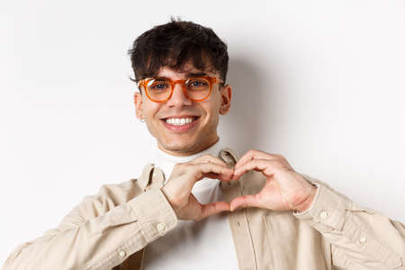 Close-up of young natural guy in glasses smiling, showing heart love you gesture, standing on white backgroundの写真素材
