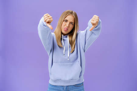Indoor shot of moodie displeased and unimpressed cool modern female with blond hair and tanned skin tilting head sticking out tongue showing thumbs down in dissatisfaction posing against purple wallの写真素材