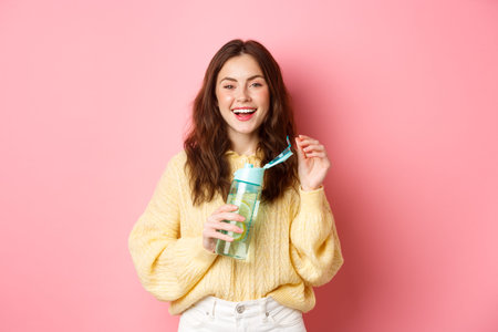 Portrait of happy brunette woman with curly hairdo, drinking water with lemon from personal bottle,...