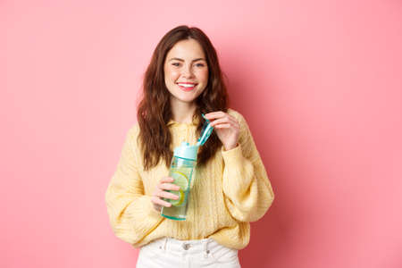 Young attractive and healthy woman smiling at camera, open water bottle with lemon, drinking sport drink, standing in colorful clothes against pink backgroundの写真素材