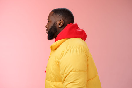 Profile studio shot bearded young 25s african guy in yellow jacket red hoodie look left normal unbothered relaxed expression standing queue order fastfood pink background, waiting take-awayの写真素材