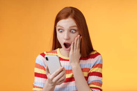 Portrait shocked concerned young redhead sensitive impressed redhead woman stare smartphone display touch cheek drop jaw stunned surprised standing orange background hold phoneの写真素材