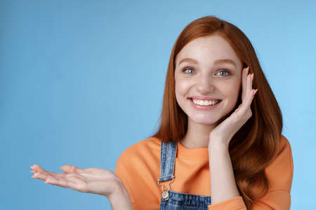 Cute tender chatismatic glad smiling redhead girl presenting awesome product show object palm hold hand raised blank copy space grinning impressed receive silly charming gift, blue backgroundの写真素材