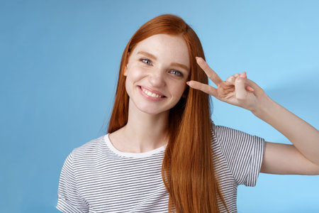 Cheerful friendly gorgeous redhead girl glancing happily show peace victory sign tilting head cute smiling broadly white teeth having fun express positivity optimism, standing blue backgroundの写真素材