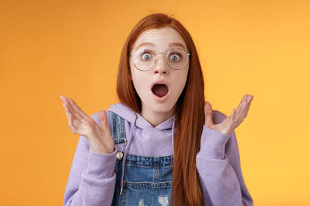 Close-up shocked sensitive concerned young panicking redhead woman worry drop jaw gasping raise hands spread freak out stare surprised emotional reacting incredible news, orange backgroundの写真素材