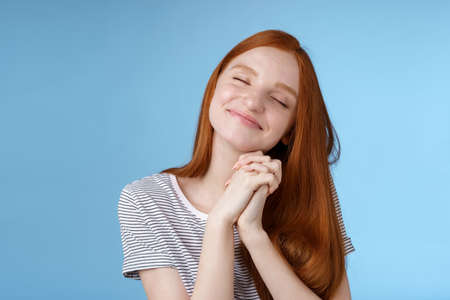 Charming delighted happy dreamy redhead girlfriend ginger long hair tilting head close eyes smiling pleased daydreaming thinking about sweet tender memories press palms together, blue backgroundの写真素材