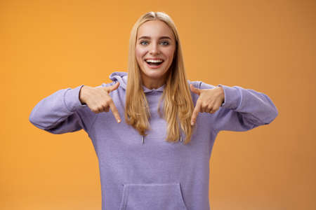 Amused joyful nice blond girlfriend pointing down present cool new product smiling broadly recommending try check out standing orange background happily grinning in hoodieの写真素材
