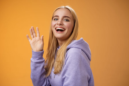 Waist-up friendly lucky cute blond girl waving palm happily smiling turning camera greet friend say hello hi see familiar person welcoming newcomers joyfully, standing orange backgroundの写真素材