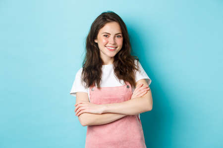 Happy smiling girl with cute blush, wearing summer clothes and beaming from joy, holding arms crossed over chest, standing against blue backgroundの写真素材