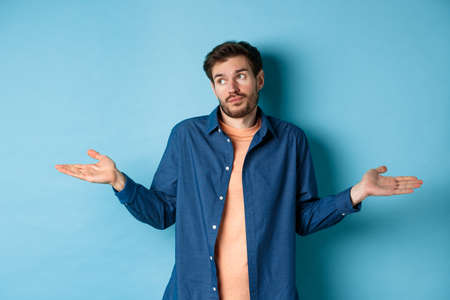 Indecisive young man with beard, look away and shrugging shoulders, know nothing, being clueless, standing on blue backgroundの写真素材