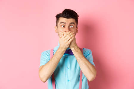 Funny young man covering mouth with hands and squint eyes, making clown grimaces, standing silly on pink background, entertain peopleの写真素材