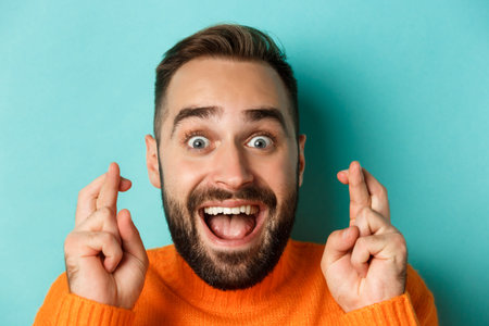 Headshot of hopeful bearded man making a wish, smiling and holding fingers crossed for good luck,...