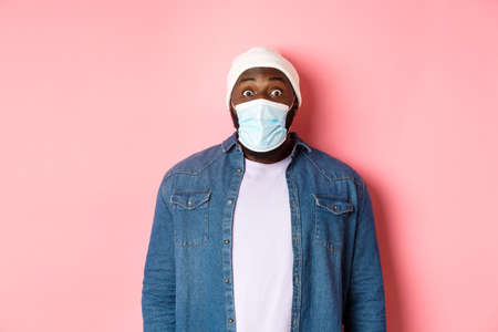Covid-19, lifestyle and quarantine concept. Image of impressed african-american man in face mask staring at camera, gasping surprised, standing over pink backgroundの写真素材