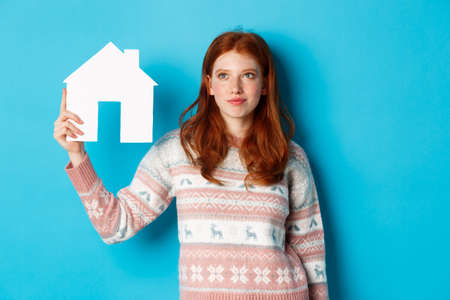 Real estate concept. Image of cute redhead girl looking curious at paper house model, thinking of buying property, looking up at copy space, blue backgroundの写真素材