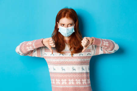 Winter, covid-19 and pandemic concept. Moody redhead girl in face mask showing thumbs-down, disapprove and dislike, standing against blue backgroundの写真素材