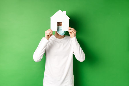 Real estate and covid-19 pandemic concept. Funny young man in face mask and white long-sleeve hiding...