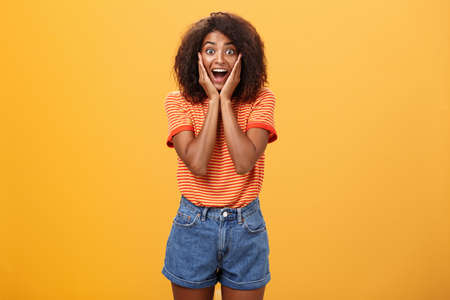 Impressed talkative african american woman finding out hot details of rumor being excited, thrilled leaning face on palms, smiling broadly with amused expression listening carefully over orange wallの写真素材
