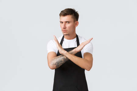 Employees, grocery stores and coffee shop concept. Serious-looking disappointed barista, waiter or cafe staff in black apron, making cross sign to show stop, warning or disapproval of actionの写真素材