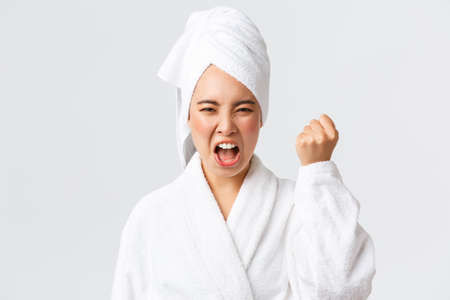 Personal care, women beauty, bath and shower concept. Close-up of mad young asian woman in bath towel and bathrobe shaking fist, scolding someone angry, threaten person, white backgroundの写真素材