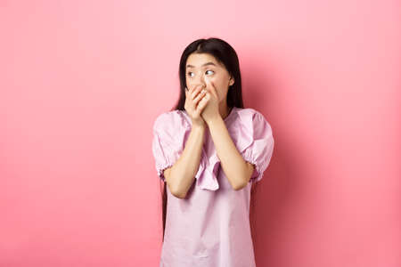 Shocked and startled asian woman looking aside at logo, covering mouth with hands speechless, standing in dress on pink backgroundの写真素材