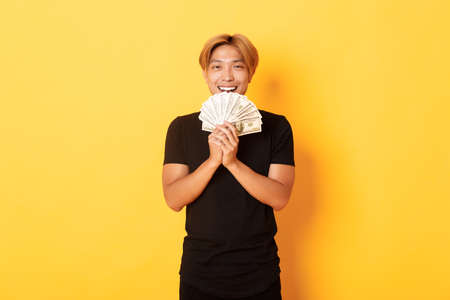 Cheerful winning handsome asian guy, smiling pleased and holding money, feeling lucky, standing yellow backgroundの写真素材