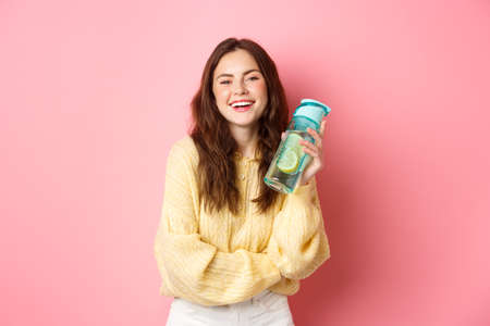 Stay hydrated. Smiling healthy girl holding water bottle with lemon and looking cheerful at camera, standing against pink studio backgroundの写真素材