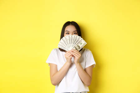 Cute asian woman hiding face behind money, peeking at camera satisfied, earn cash, standing over yellow backgroundの写真素材
