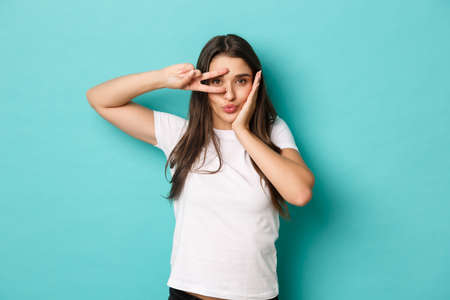 Image of silly glamour girl in white t-shirt, pouting and showing peace sign over eye, standing flirty over blue backgroundの写真素材