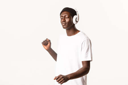 Portrait of carefree attractive african-american guy, close eyes and dancing while listening music in wireless headphones, white backgroundの写真素材