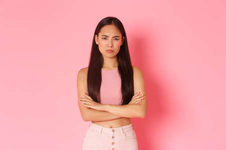Lifestyle, beauty and women concept. Waist-up portrait of angry and offended cute sulking asian girlfriend cross arms, frowning and looking displeased, mad at someone, pink backgroundの写真素材